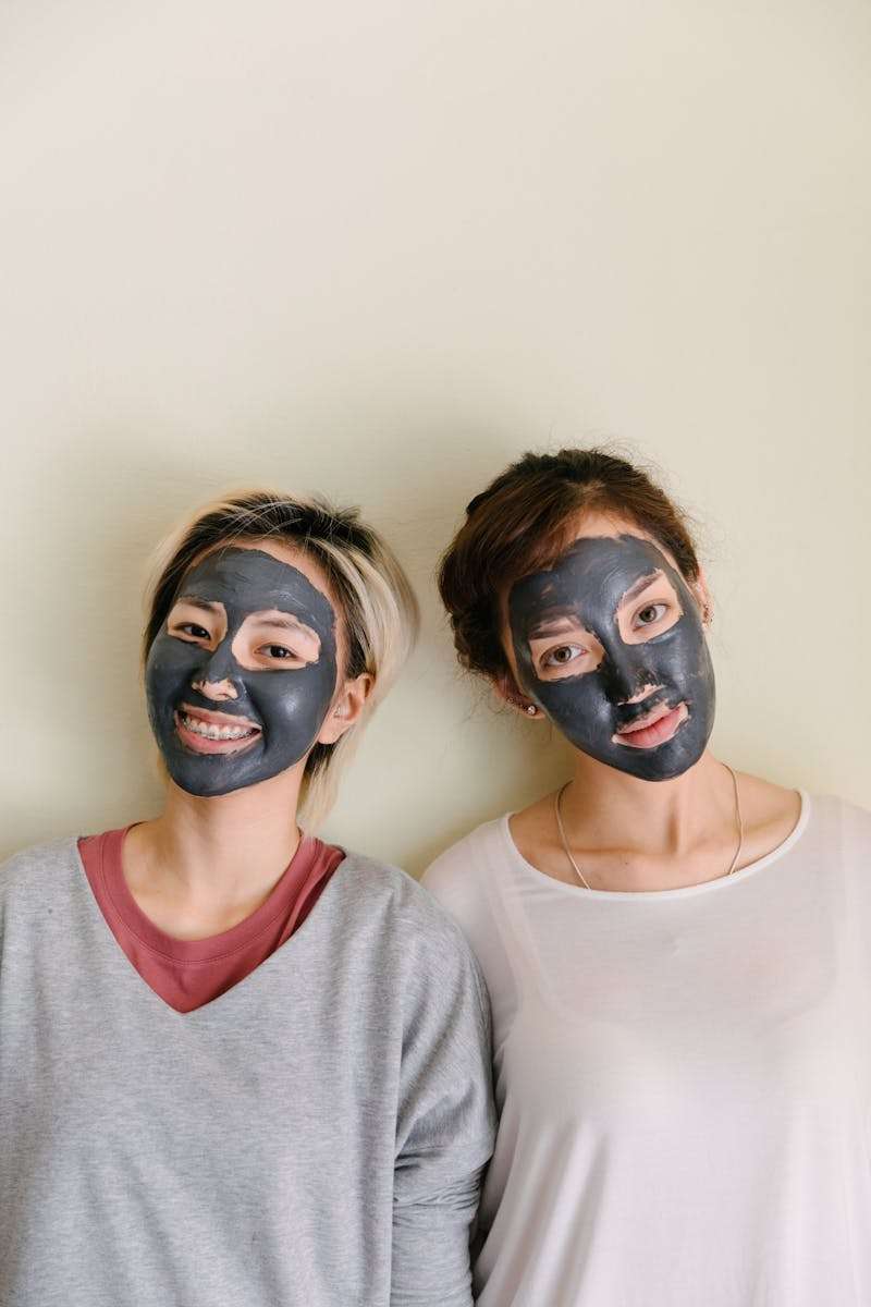 Young diverse women in casual clothes with black facial mud mask on beige background looking at camera